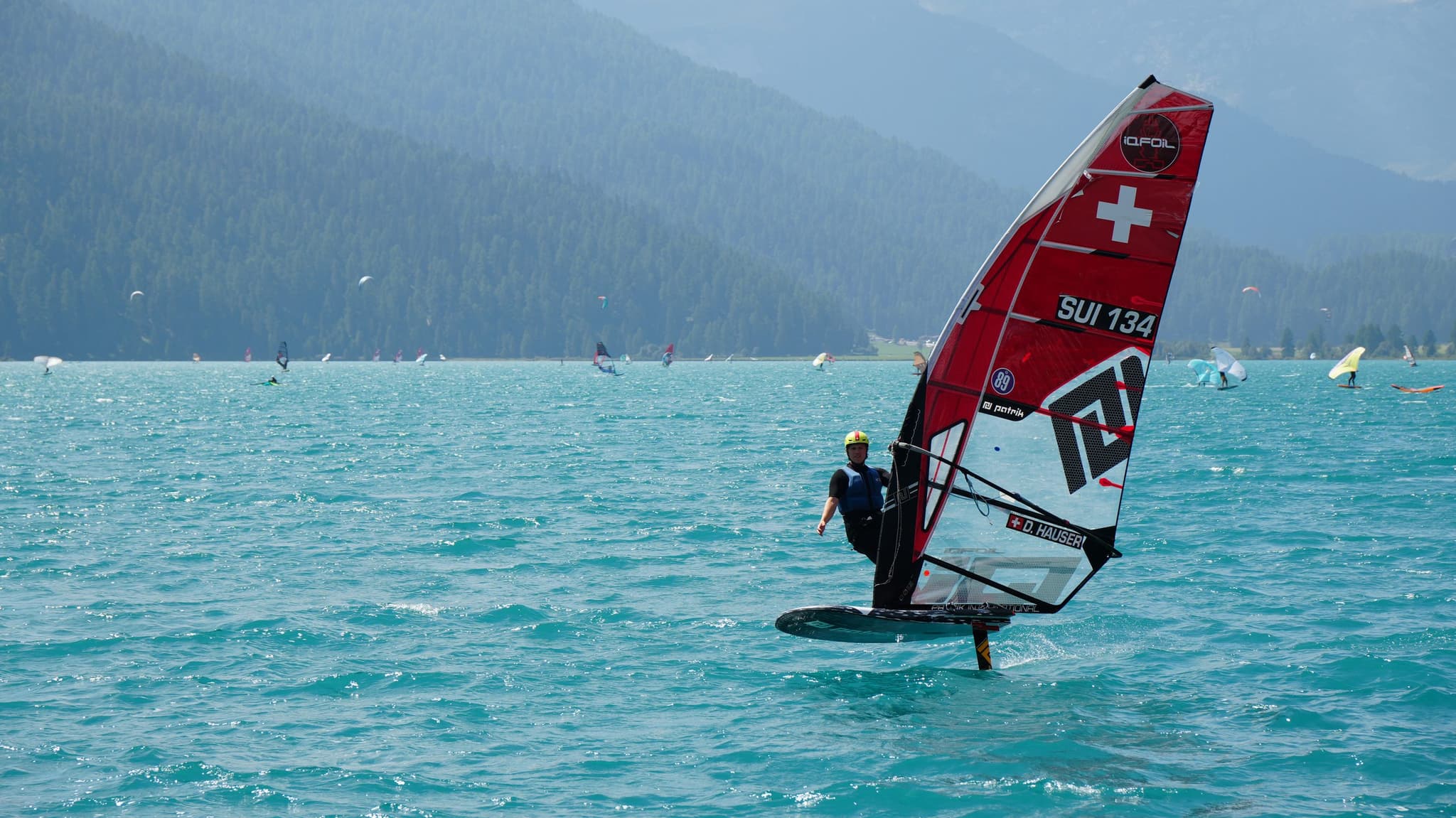 Devin Hauser foilt auf dem Silvaplanersee, rotes iQFOiL-Segel mit Schweizerkreuz und Segelnummer SUI-134 vor Bergkulisse